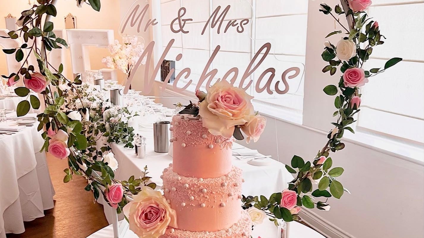 A blush pink tiered wedding cake decorated with piped pearls and fresh roses, beside a floral hoop at a wedding reception